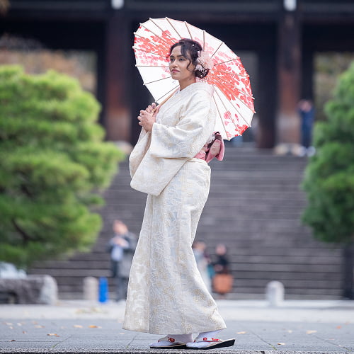 A foreign traveler wearing an elegant Kimono and holding a traditional paper umbrella in front of a historic shrine in Kyoto.