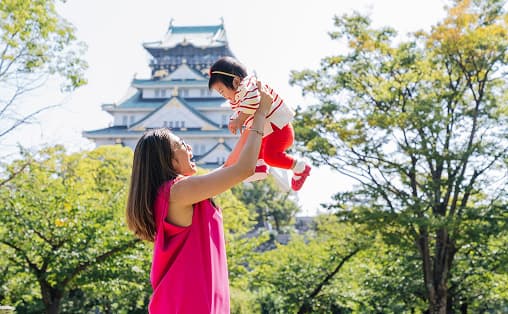 A foreign mother joyfully lifting her baby high in a lush, green Japanese park during a relaxed family photo session.